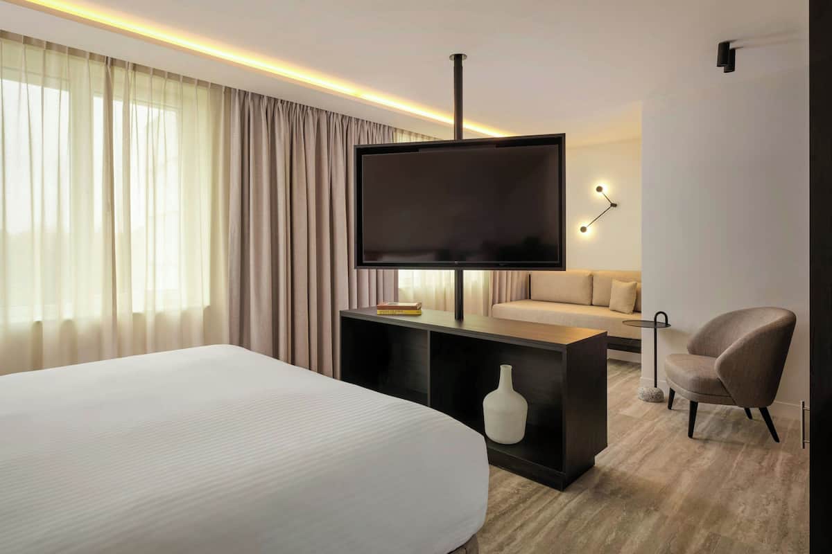 A modern hotel room with a large bed, a flat-screen TV, a sofa, a chair, and a small table.