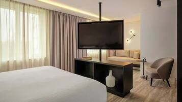 A modern hotel room with a large bed, a flat-screen TV, a sofa, a chair, and a small table.