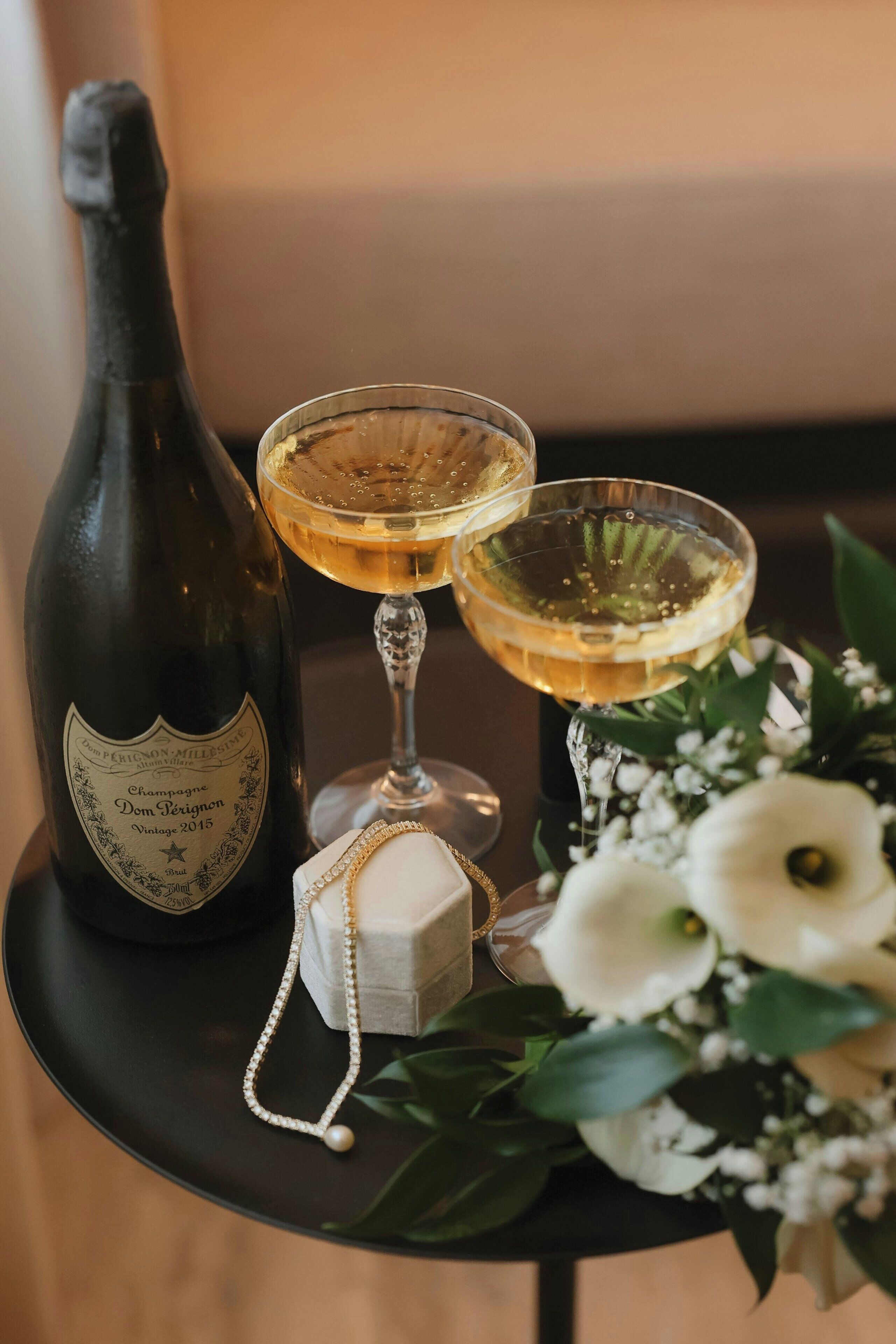 A bottle of champagne, two glasses of champagne, a small box, and a flower arrangement on a table.