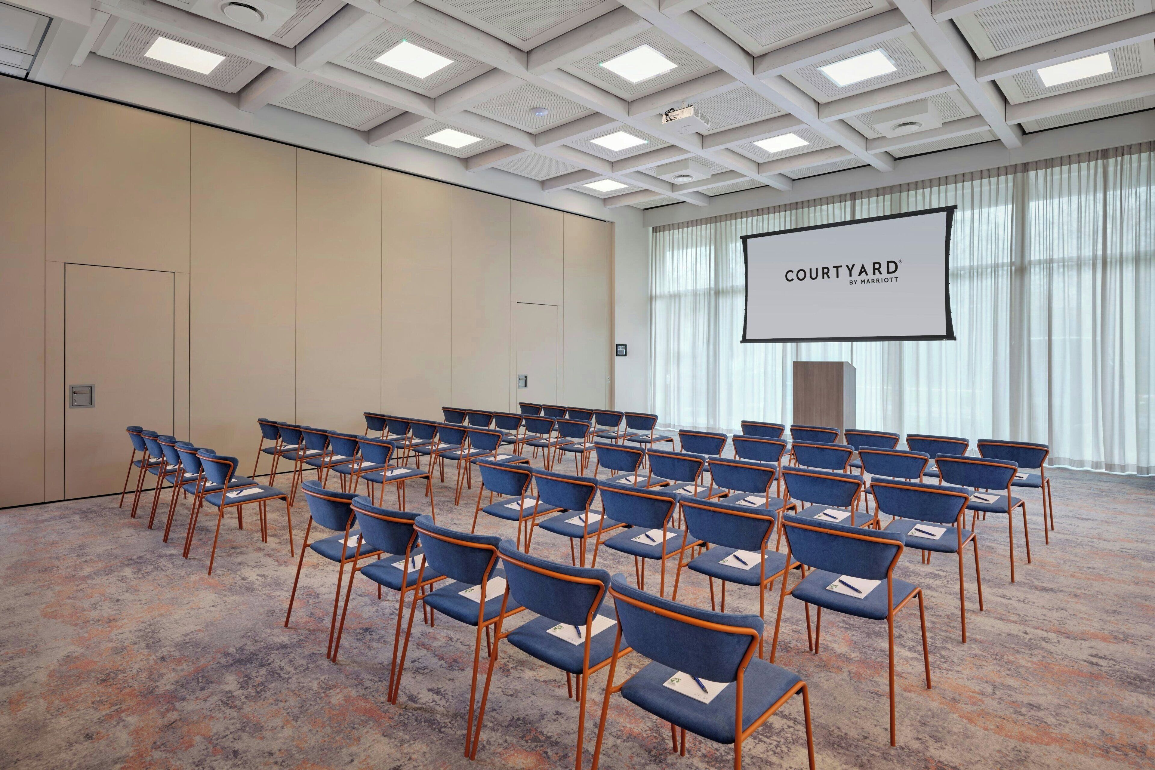 Ein Konferenzraum mit blauen Sitzreihen, einem großen Bildschirm, der 'COURTYARD BY MARRIOTT' zeigt, und einem Teppichboden.