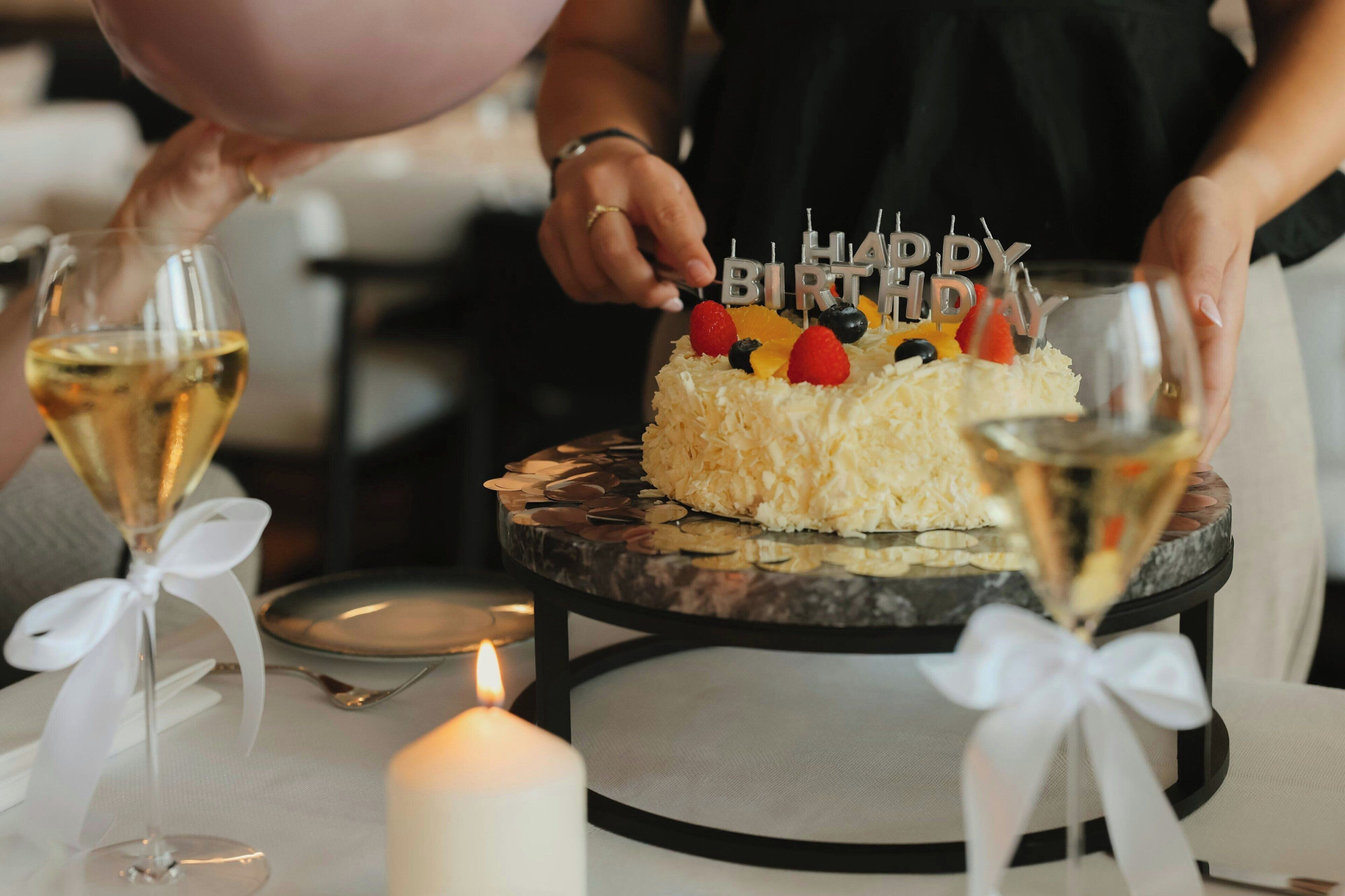 Una torta di compleanno con candeline che riportano la scritta 'HAPPY BIRTHDAY' su un tavolo con due bicchieri di vino bianco e una candela accesa.