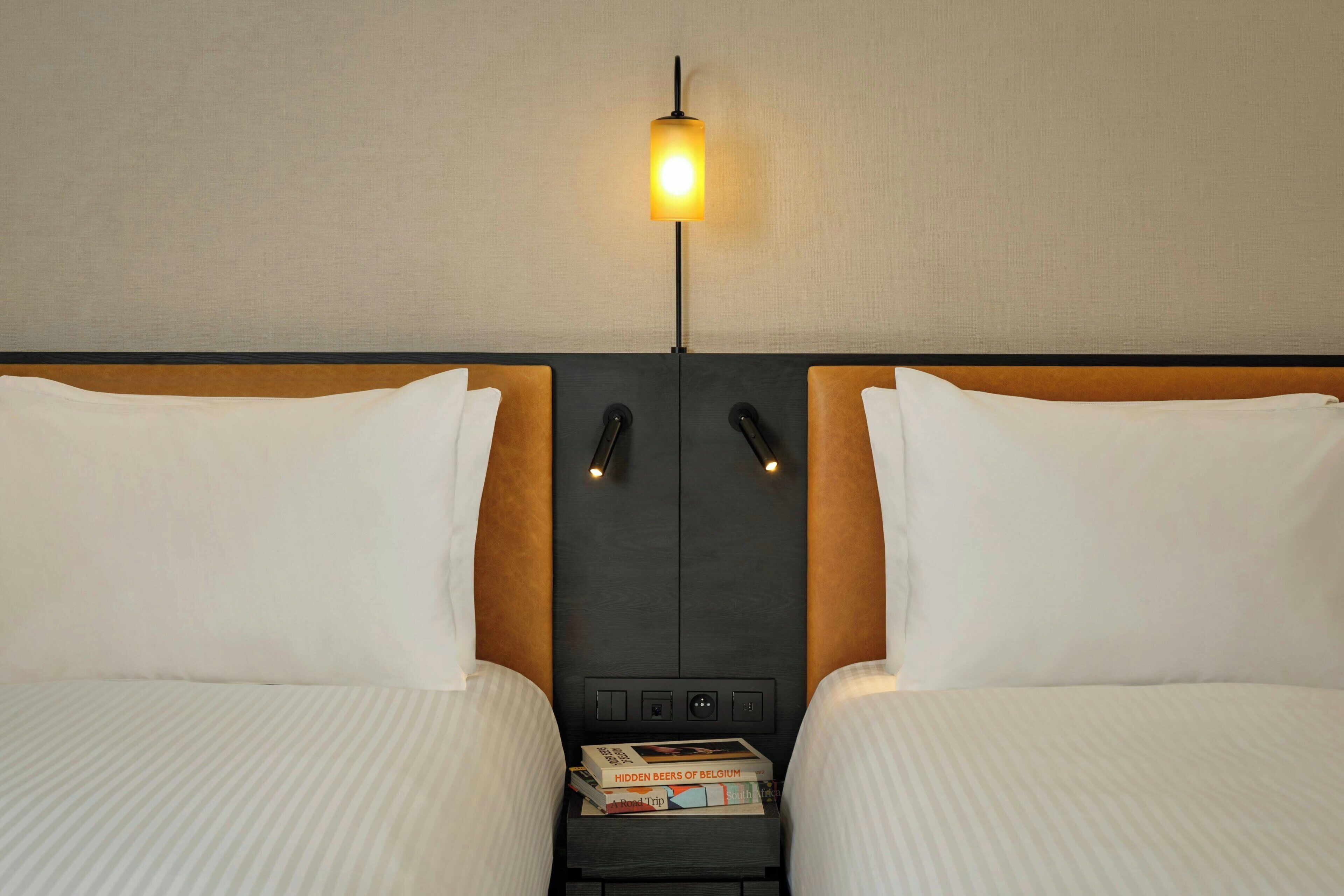 Ein Hotelzimmer mit zwei Betten, einem Nachttisch mit Büchern, einer Wandleuchte und einer Bedienkonsole.