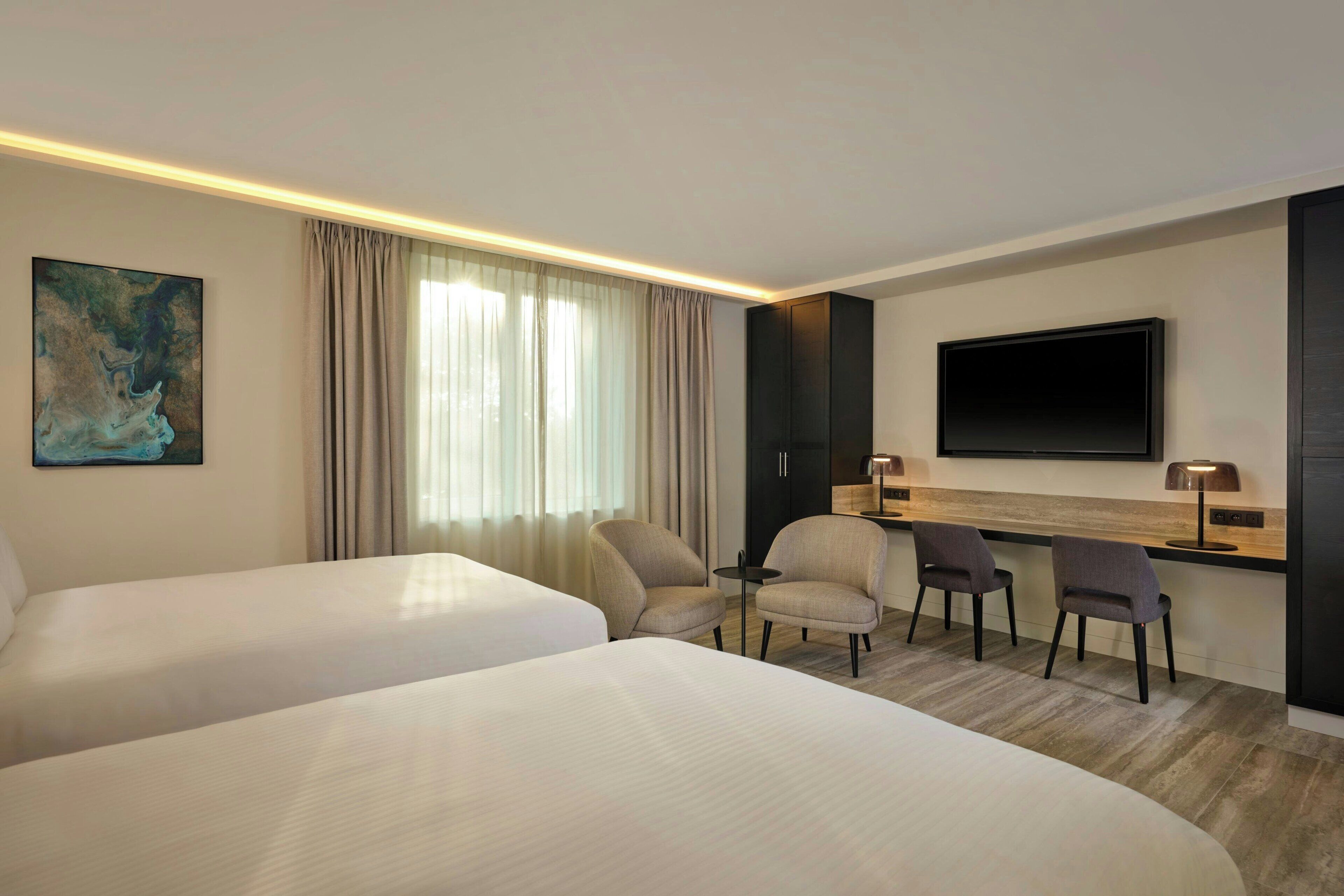 A hotel room with two beds, a desk with chairs, a flat-screen TV, and a painting on the wall.