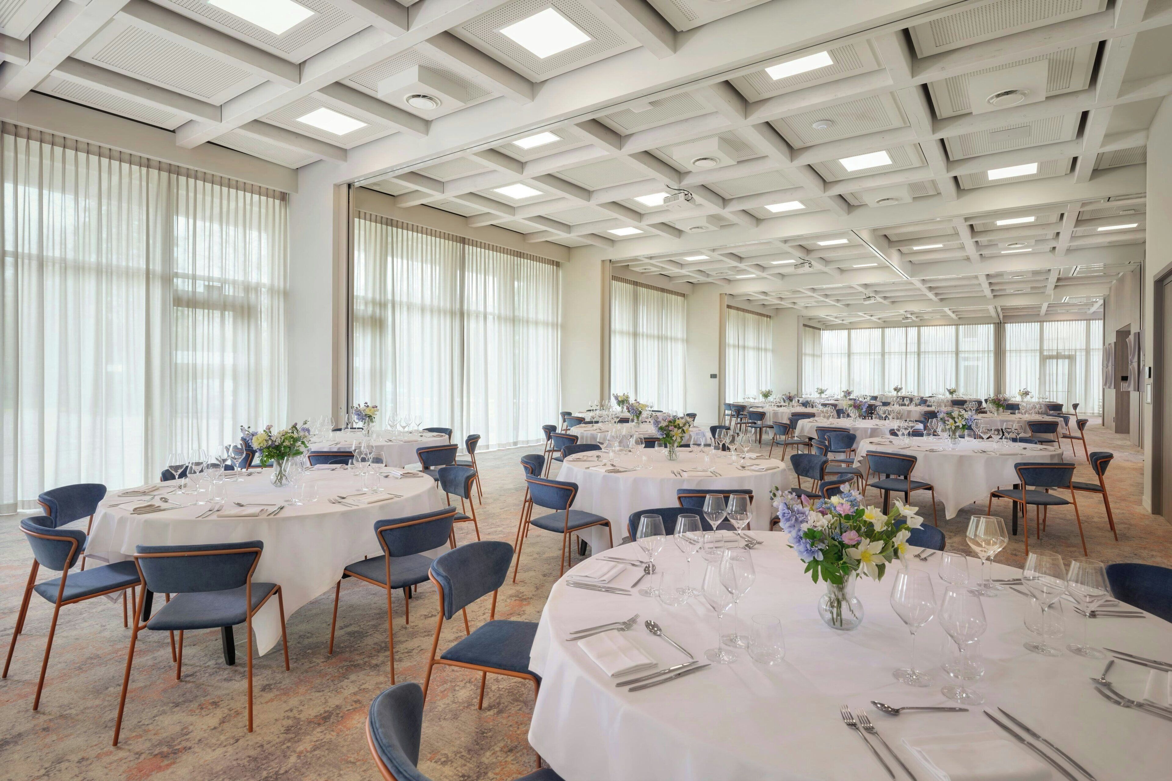 A large banquet hall with multiple round tables set for dining, featuring white tablecloths and blue chairs.