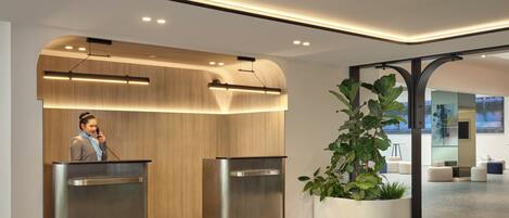 Una moderna zona de recepción con un panel de madera, una planta grande en maceta y una persona en un atril.