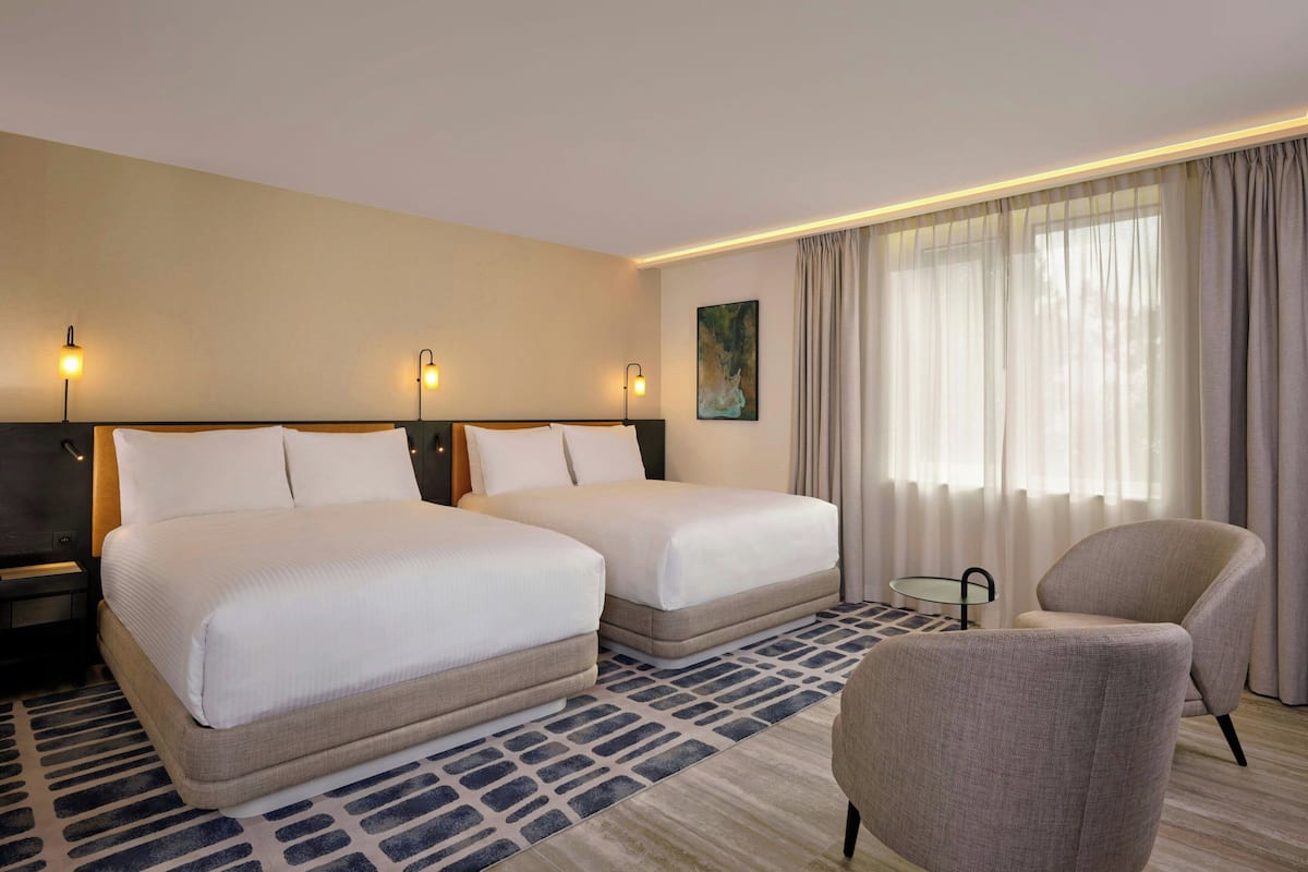 A hotel room with two beds, a chair, a small table, and a painting on the wall.