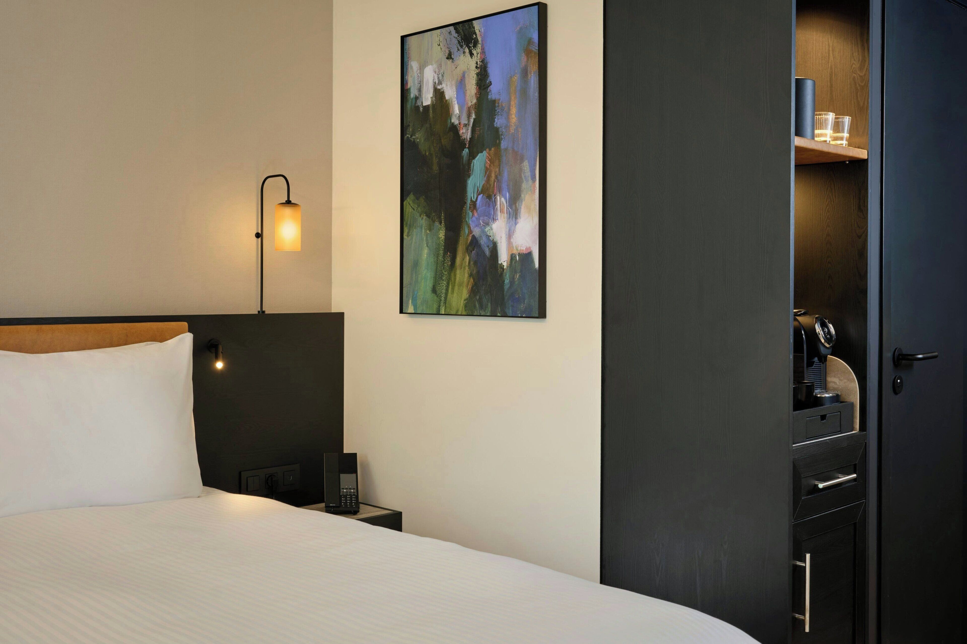 Ein Hotelzimmer mit einem Bett, einer Nachttischlampe, einem Telefon, einem Bild an der Wand und einem Kleiderschrank.