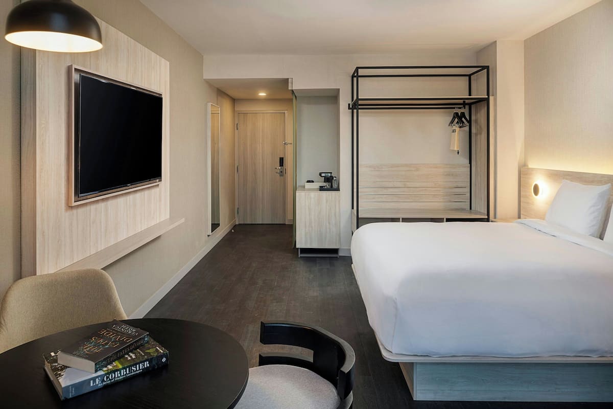 A hotel room with a bed, a TV mounted on the wall, a desk with a chair, and a small table with books.