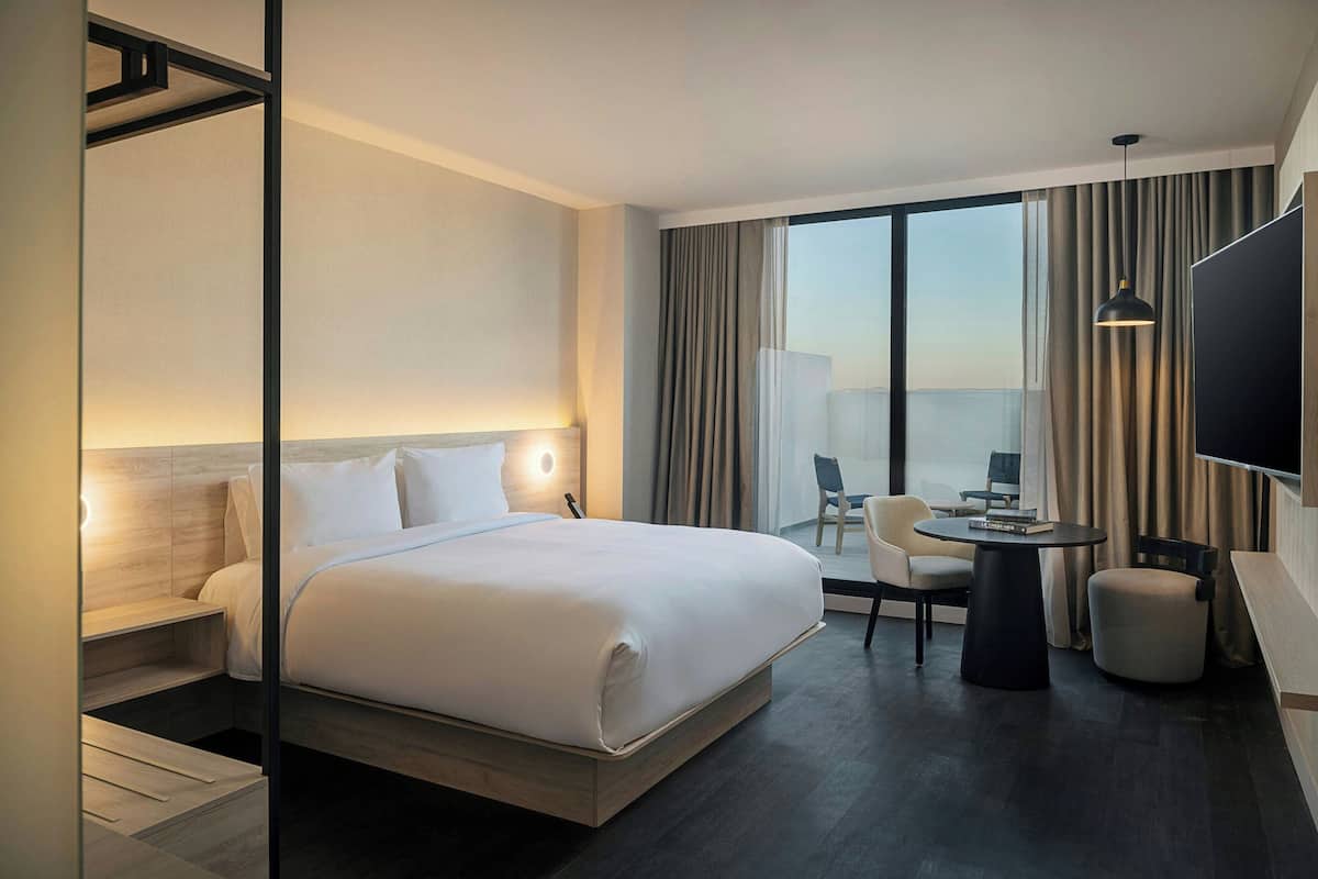 A modern hotel room with a large bed, a desk with a chair, a small table, and a TV.
