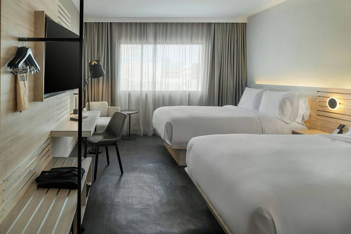 A hotel room with two beds, a desk, a chair, a TV, and a window with curtains.