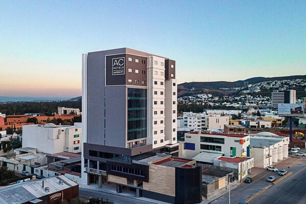 Un hotel moderno con el logotipo destacado de AC, rodeado de otros edificios y un cielo despejado.