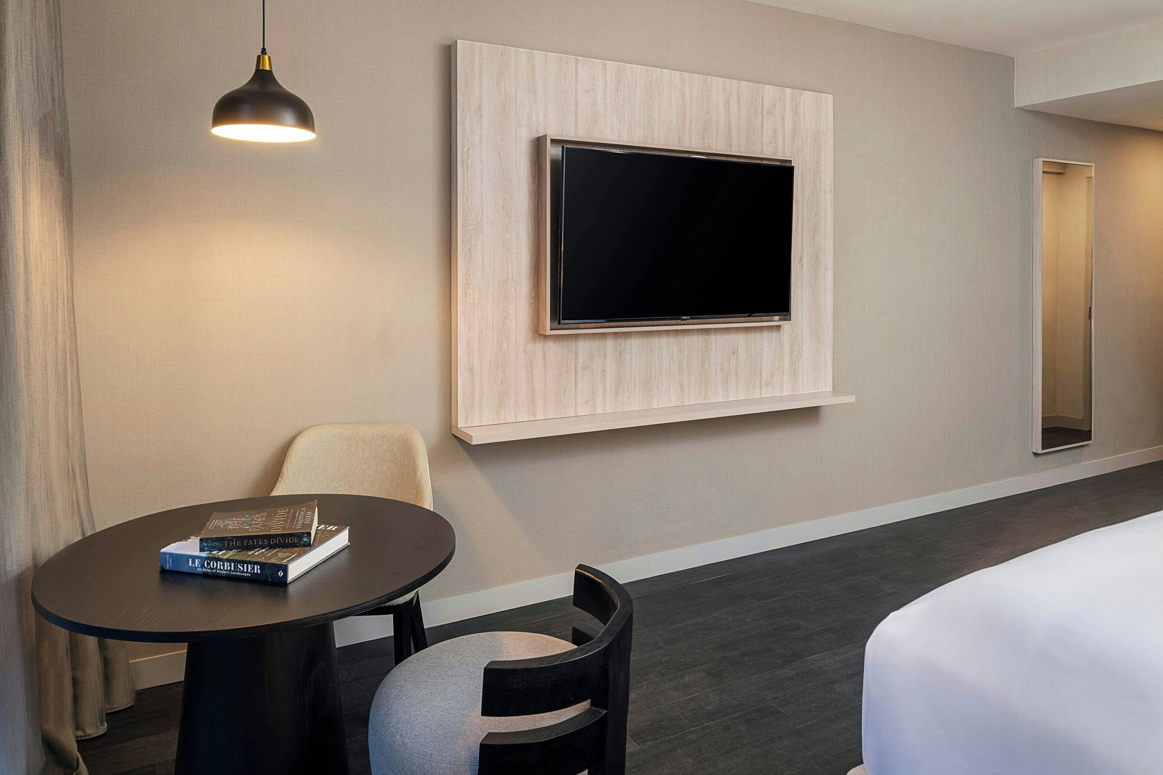 Habitación de hotel con una televisión de pantalla plana montada en la pared, una mesa redonda con dos sillas y una cama con edredón blanco.