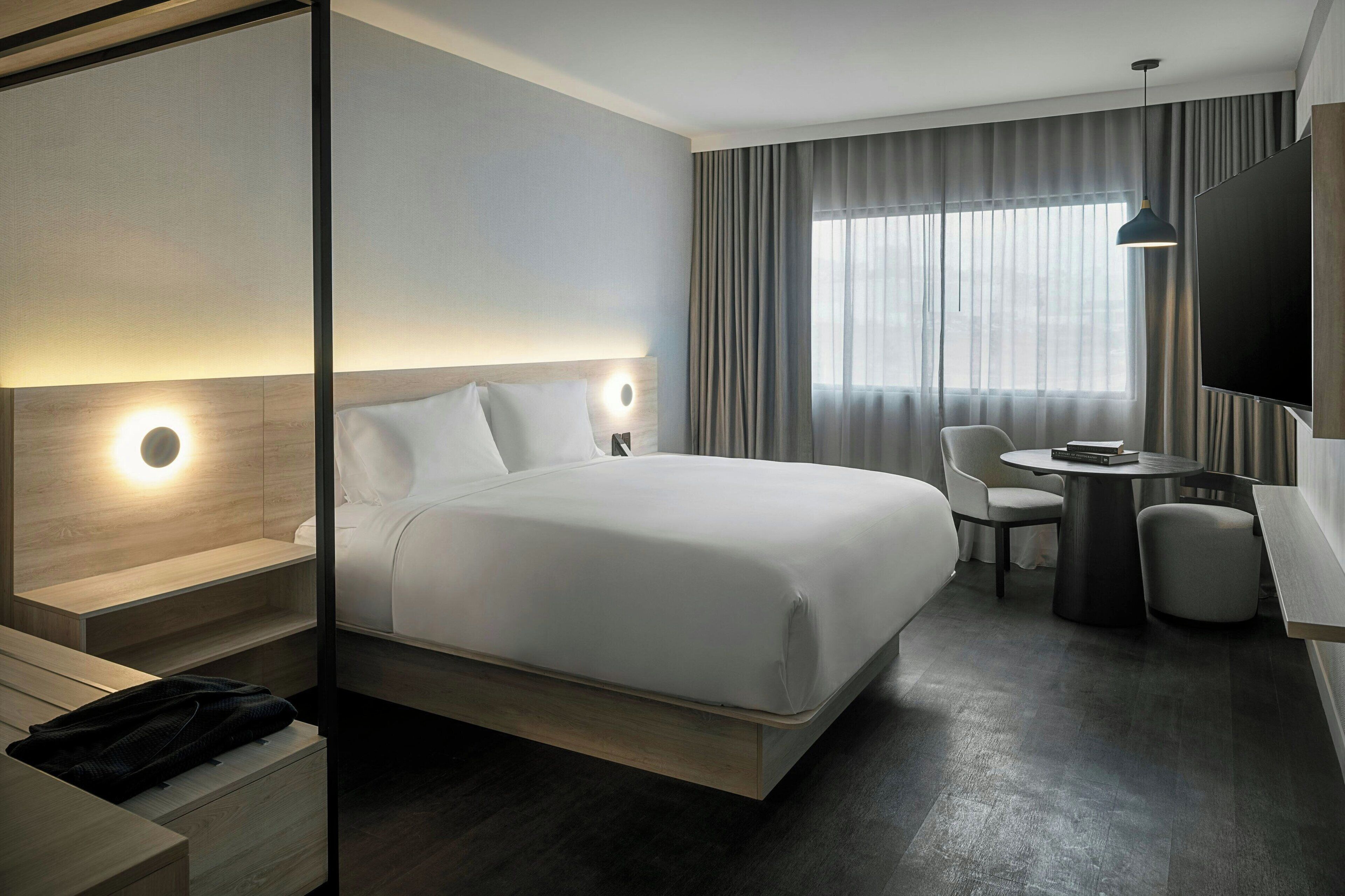 Ein modernes Hotelzimmer mit einem großen Bett, Nachttischen, einem Schreibtisch mit Stuhl, einem runden Tisch und einem Fernseher.