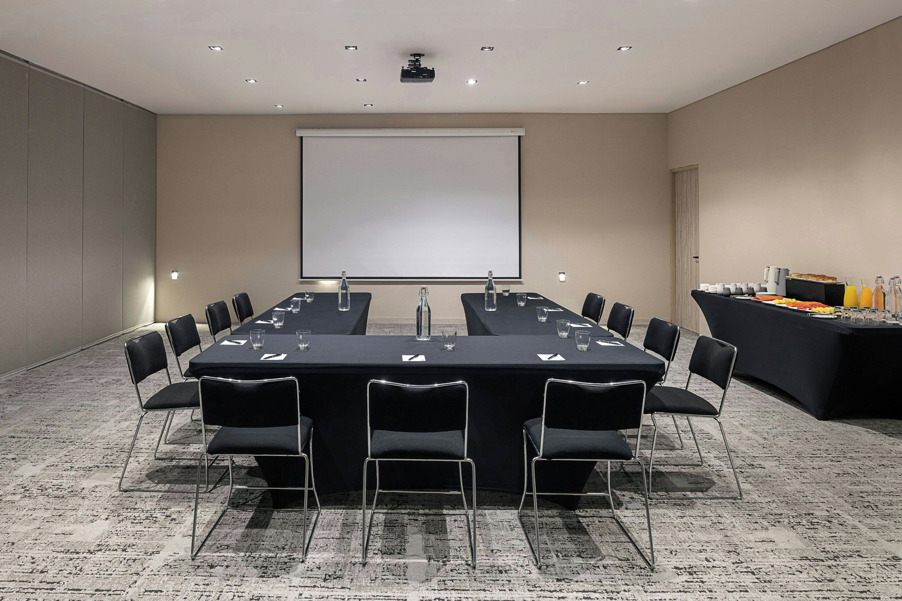Un salón de conferencias con una larga mesa, sillas, una pantalla de proyector y una mesa de buffet.