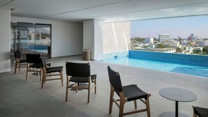 A modern indoor pool area with a view of the city, furnished with wooden chairs and a round table. - AC Hotel By Marriott San Luis Potosi (San Luis Potosi)