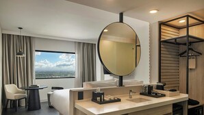A modern hotel room with a large bed, a round mirror, a vanity with a sink, a small table, a chair, and a view of the city through the window. - AC Hotel By Marriott San Luis Potosi (San Luis Potosi)