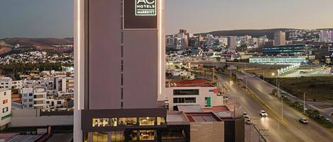 Un hotel moderno con el logo de AC destacado, una fachada bien iluminada y vistas a la ciudad de fondo.