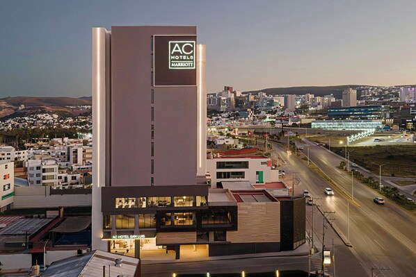 Un hotel moderno con el logo de AC destacado, una fachada bien iluminada y vistas a la ciudad de fondo.