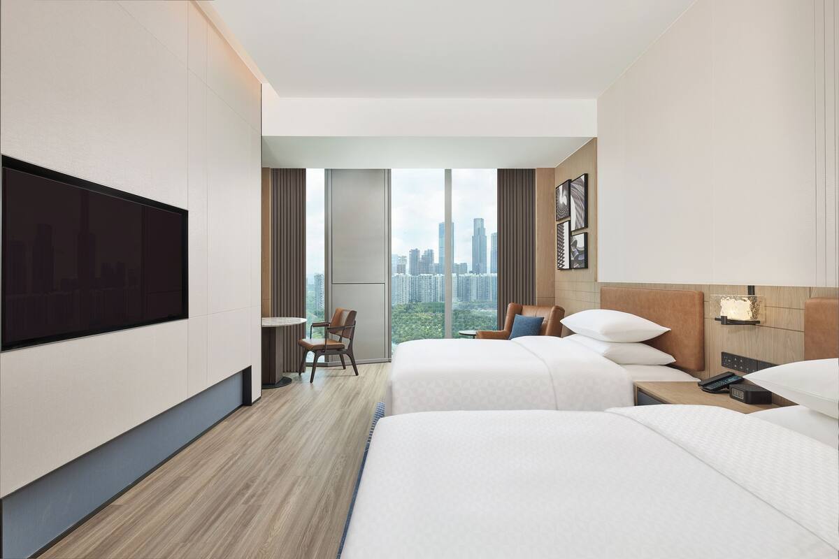 A hotel room with two beds, a TV, a desk, and a city view.