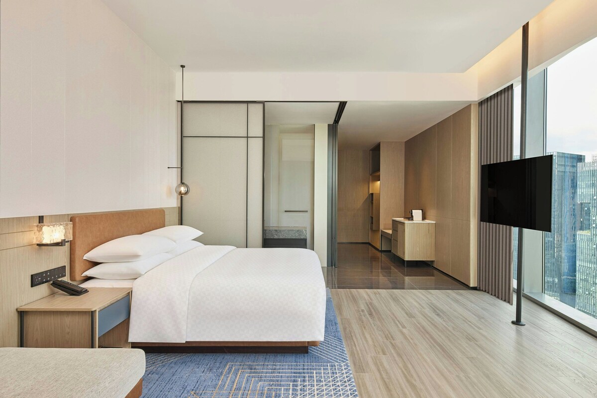 A modern hotel room with a large bed, a TV, a desk with a lamp, and a city view.