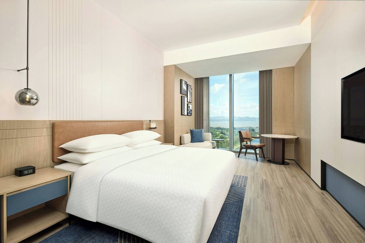 A modern hotel room with a large bed, a desk, a chair, a small table, and a TV.