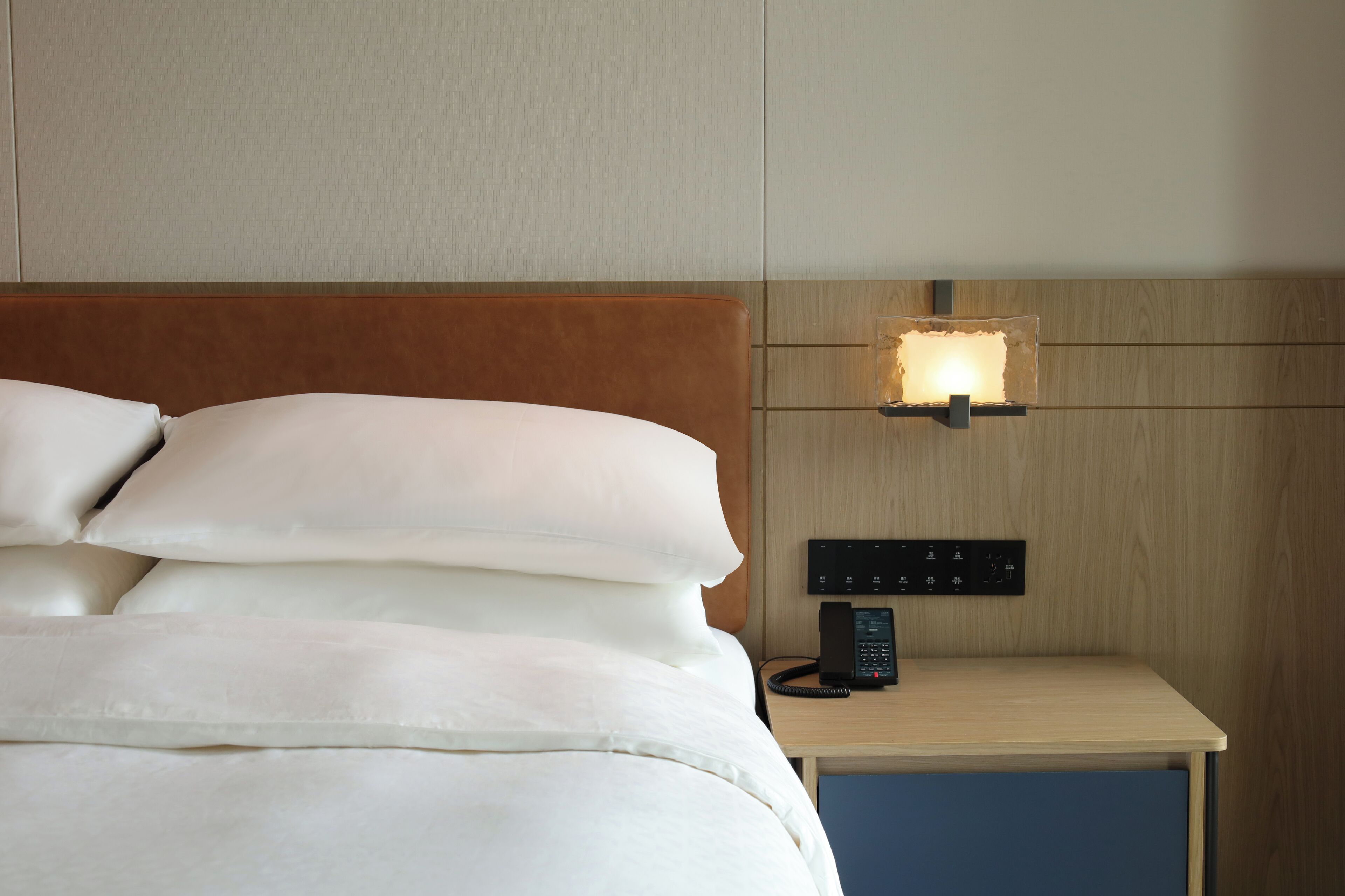 A neatly made bed with white linens, a wooden headboard, a bedside table with a lamp, and a control panel.