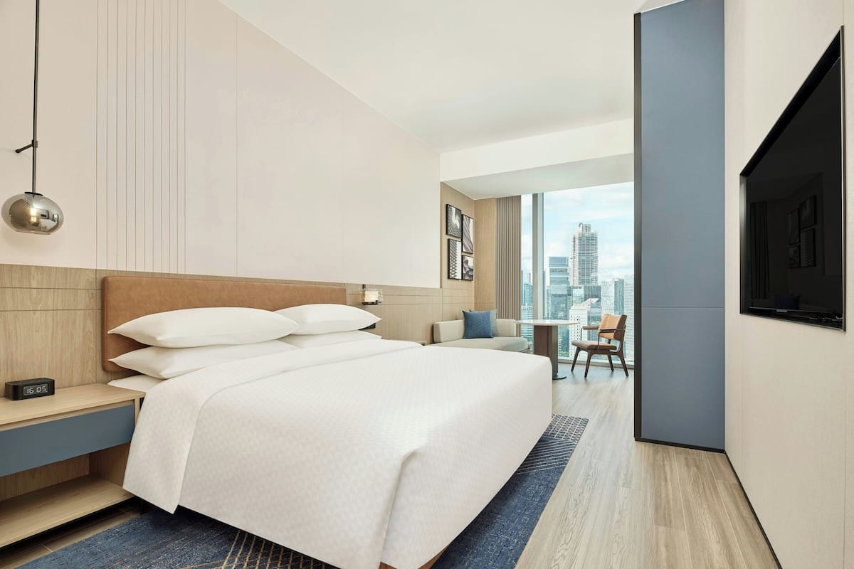 A modern hotel room with a large bed, a bedside table with a clock, a TV mounted on the wall, and a city view through the window.