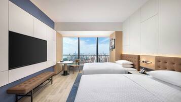 A hotel room with two beds, a bench, a TV, a desk, and a city view.