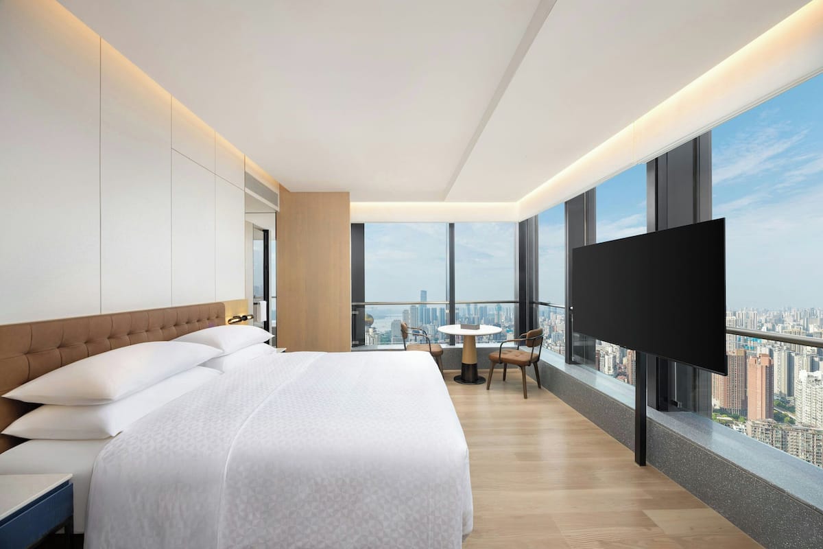 Une chambre d’hôtel moderne dotée d’un grand lit, d’une télévision et d’un balcon offrant une vue sur la ville, ainsi que d’une petite table avec des chaises.