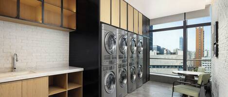 A modern laundry room with a row of washing machines, a sink, and a dining area with a table and chairs.