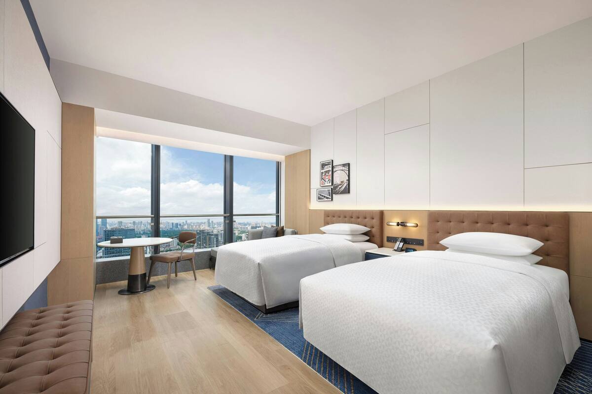 Une chambre d’hôtel moderne équipée de deux lits, d’un bureau, d’une chaise et d’une grande fenêtre donnant sur la ville.