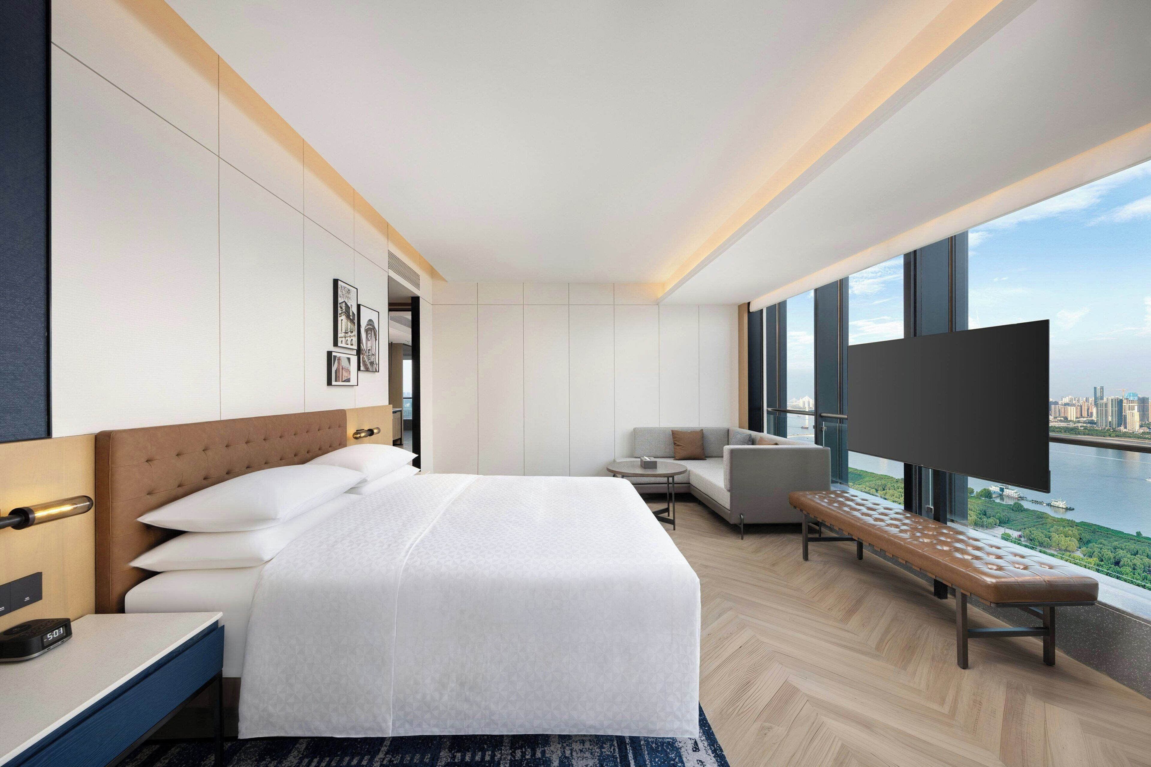 A modern hotel room with a large bed, a sofa, a TV, and a view of a river.