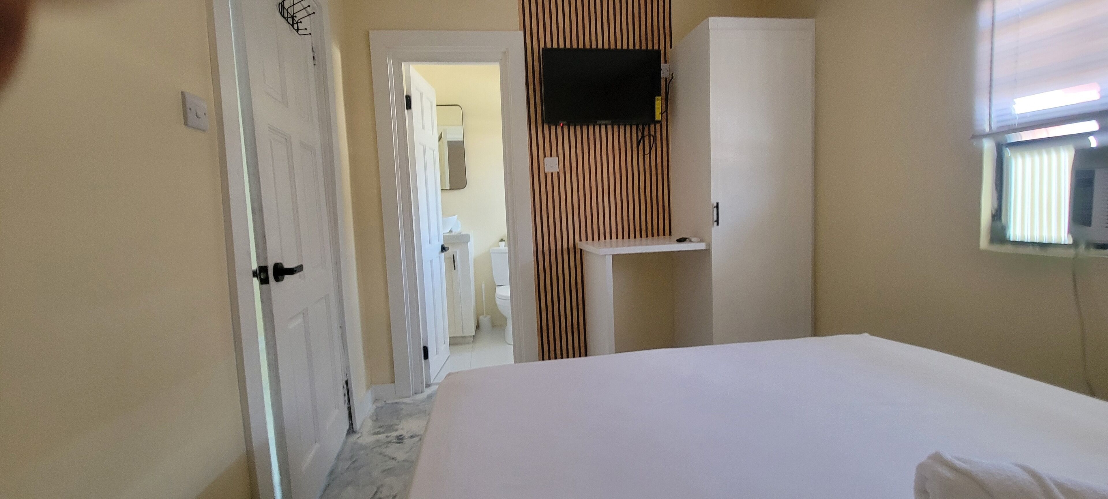 A hotel room with a bed, a television mounted on the wall, a small table, and a window with blinds.