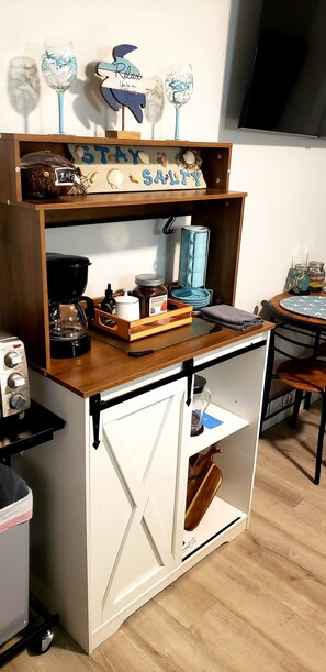 Une petite cuisine équipée d’une machine à café, d’un pot de miel et d’une étagère où sont disposés des objets décoratifs.