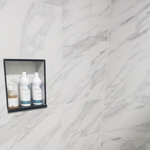 A bathroom with white marble tiles and a shelf holding three bottles of hair care products.