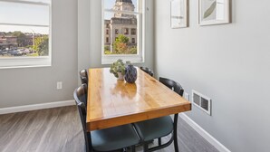 A dining area with a wooden table, black chairs, a vase with plants, and a cityscape view through a large window. - Rustic-industrial Loft With Courthouse Views 3 Bedroom Apts (Bloomington)
