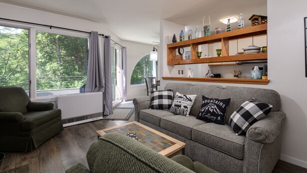 A cozy living room with a grey sofa, a green armchair, and a wooden coffee table. There is a large window with curtains, a shelf with decorative items, and a radiator. - Glades 14 1 Bedroom Condo (Warren)