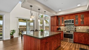 A modern kitchen with wooden cabinets, a large island, stainless steel appliances, and a view of the outdoors. - Kauai Pili Mai 14i 3 Bedroom Condo (Koloa)