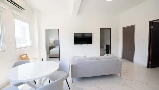 A modern living room with a white sofa, a round dining table, and a flat-screen TV mounted on the wall. - Close To The Beach Apt. 71Di.203 (San Juan)
