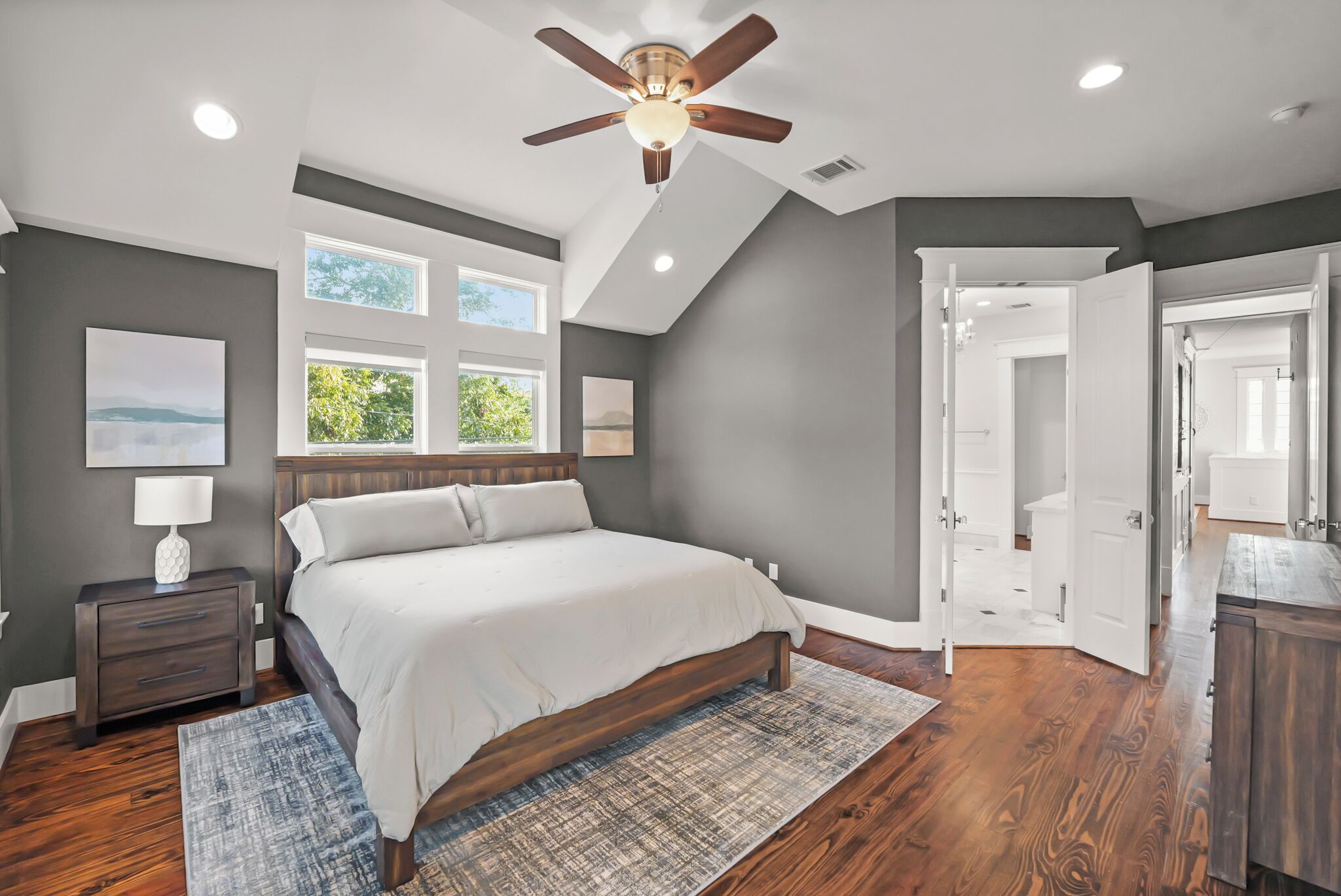 A bedroom with a large bed, a ceiling fan, and a view of the outdoors through windows.