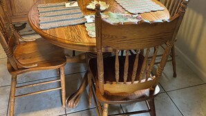 A wooden dining table with a placemat, surrounded by four wooden chairs. A stainless steel refrigerator is in the background.