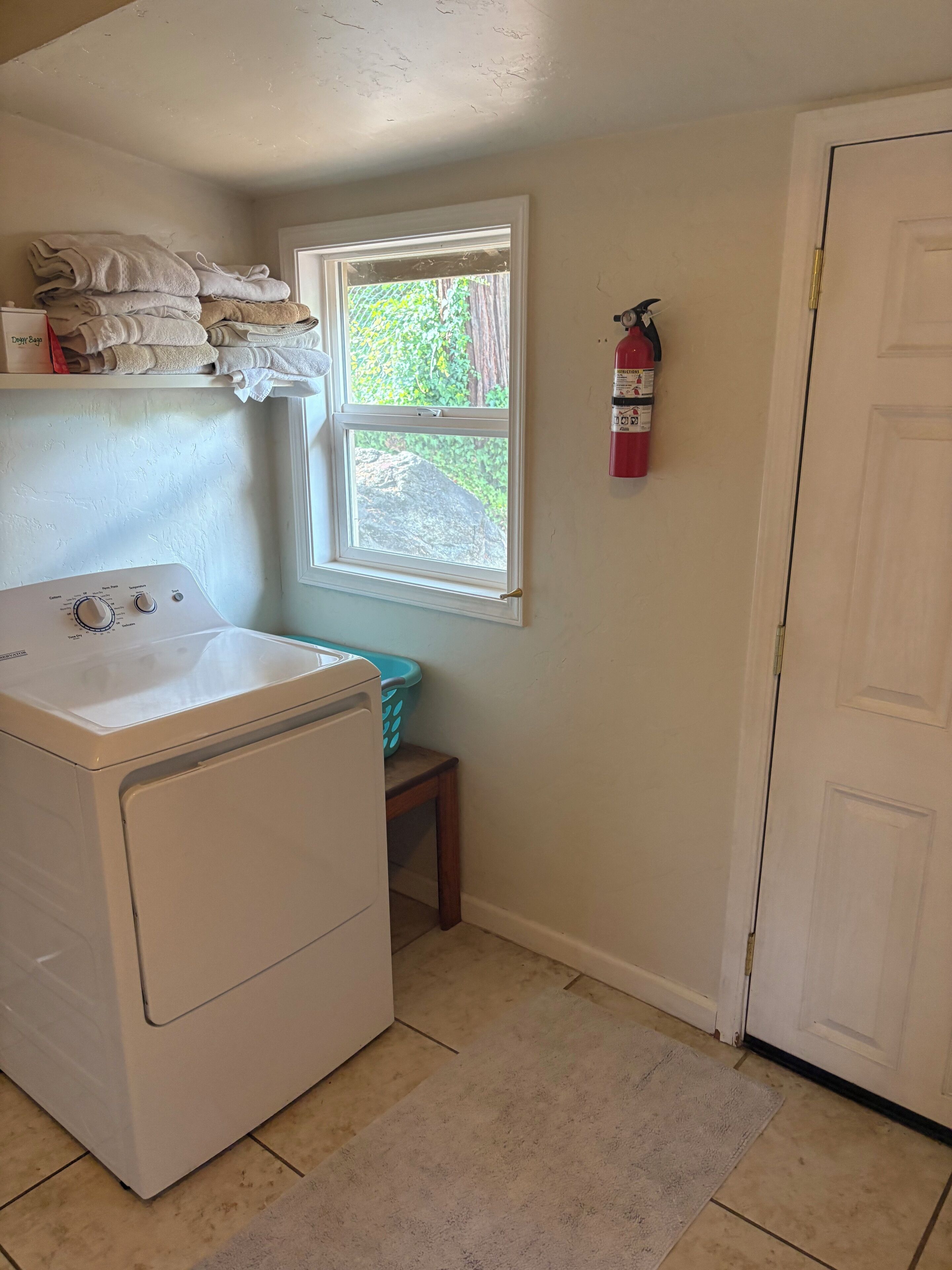 A laundry room with a front-loading washer and dryer, a window with a view of greenery, a fire extinguisher on the wall, and a door to the right.