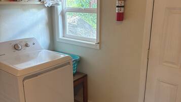 A laundry room with a front-loading washer and dryer, a window with a view of greenery, a fire extinguisher on the wall, and a door to the right.