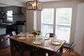 A dining area with a wooden table set for four, a kitchen in the background, and a large window with blinds. - Peaceful lake front home with amazing views. (Middlesex)