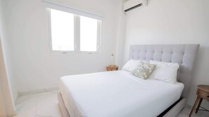 A bedroom with a large bed, a window, a bedside table, and a wall-mounted air conditioning unit.