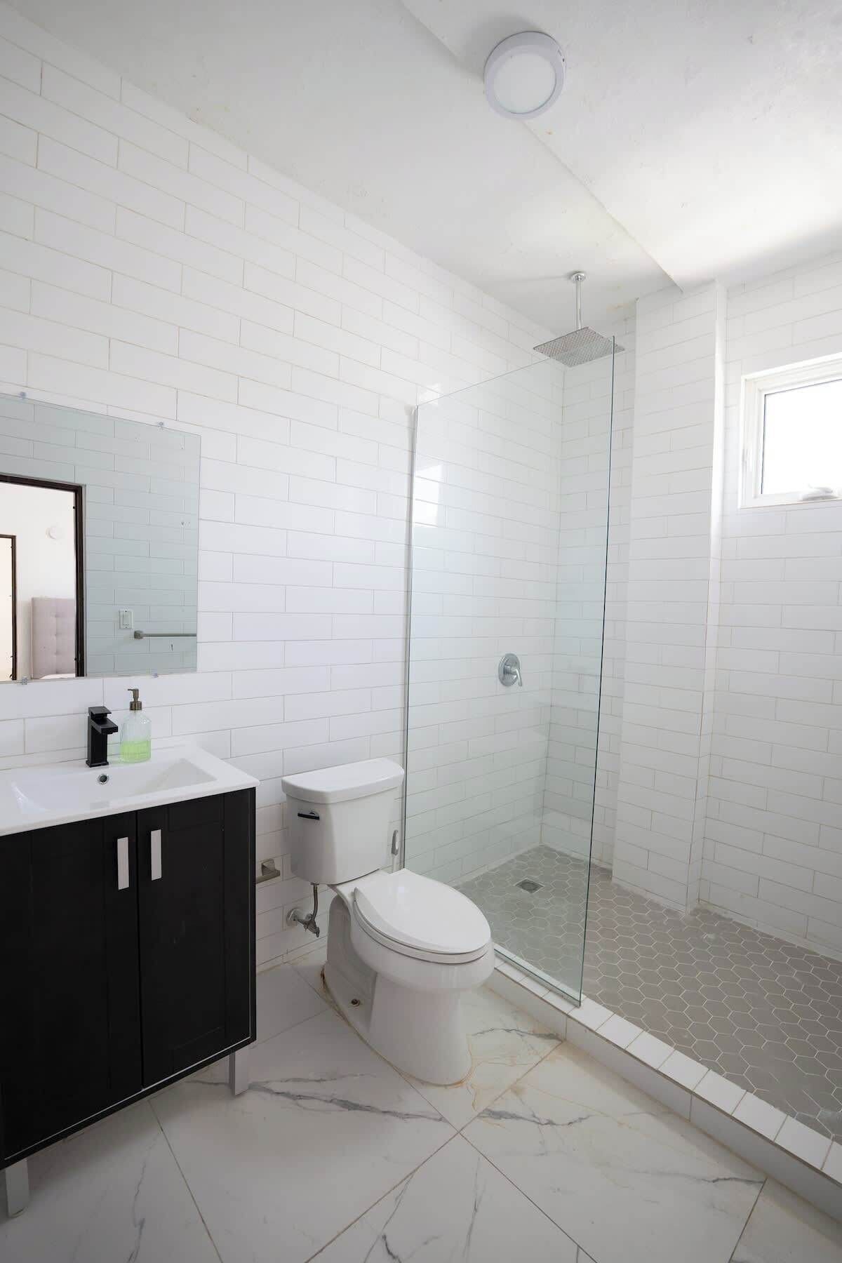 Ein modernes Badezimmer mit weißen, rechteckigen Fliesen, einer Glasdusche, einer weißen Toilette und einem schwarzen Waschtisch.