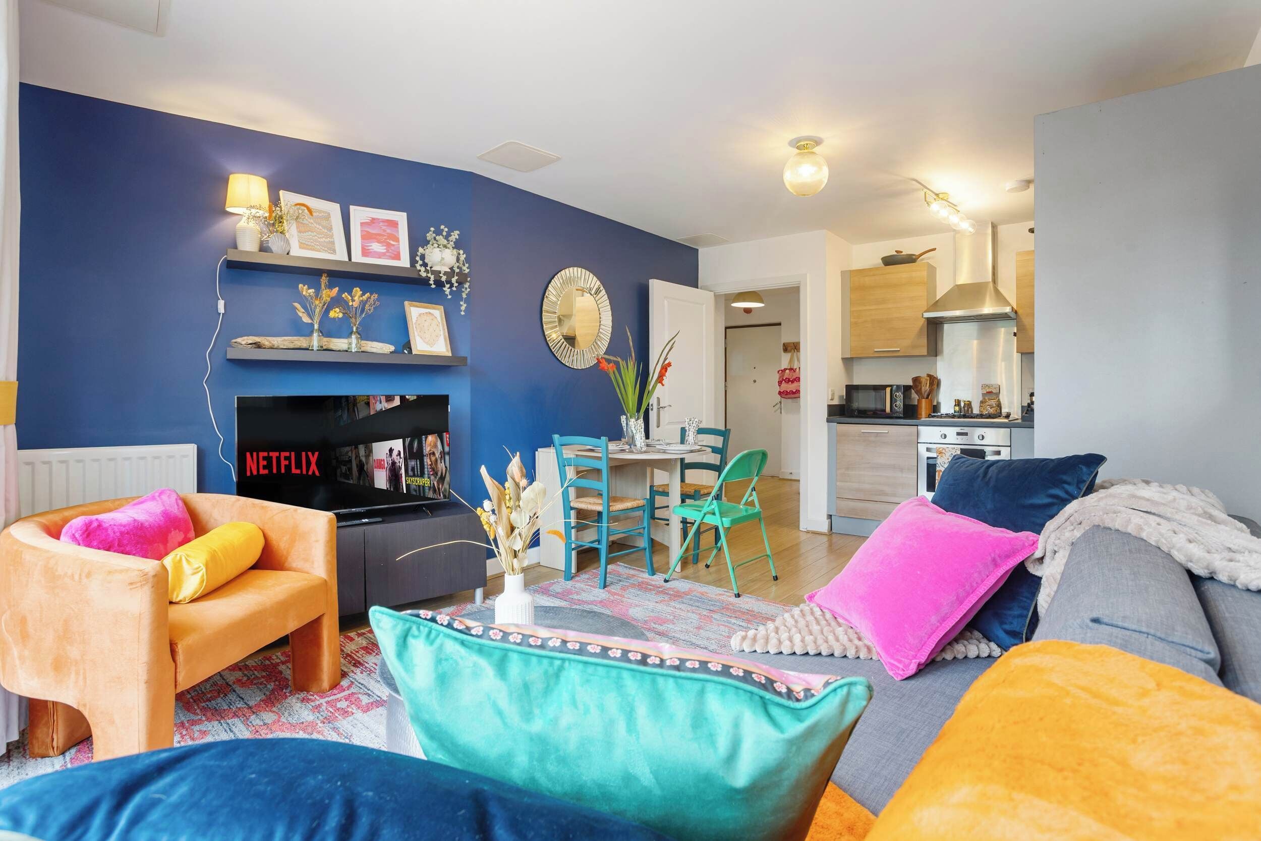 A living room with a blue wall, a TV, a fireplace, a dining area with chairs, and a kitchen in the background.