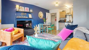A living room with a blue wall, a TV, a fireplace, a dining area with chairs, and a kitchen in the background.