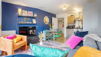 A living room with a blue wall, a TV, a fireplace, a dining area with chairs, and a kitchen in the background.