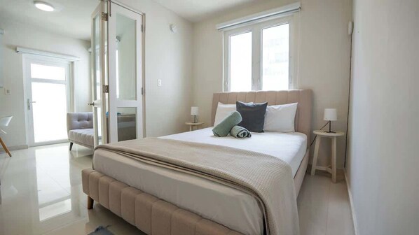 A modern bedroom with a large bed, two bedside tables with lamps, a window, and a door leading to another room.